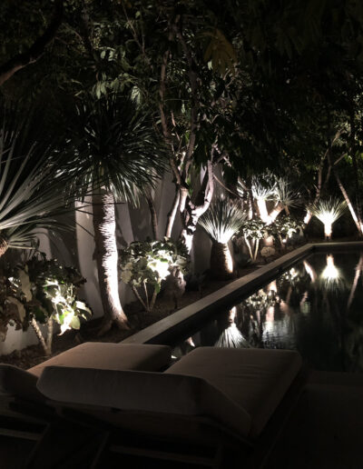 Illuminated garden pathway beside a reflective pool at night with loungers.