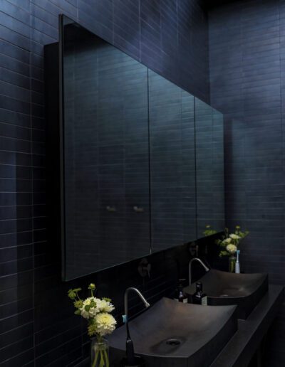Modern bathroom with dark tiles, featuring a large mirror and a double sink vanity with floral arrangements.