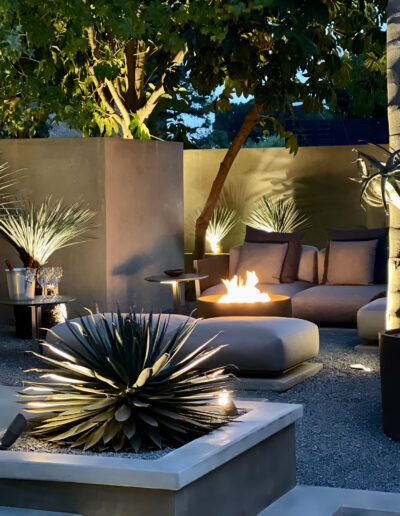 Modern outdoor patio at dusk with fire pit and comfortable seating surrounded by decorative plants.