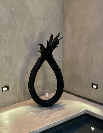 An abstract wooden sculpture resembling a loop stands next to a small body of water, illuminated by floor lights against a textured grey wall.