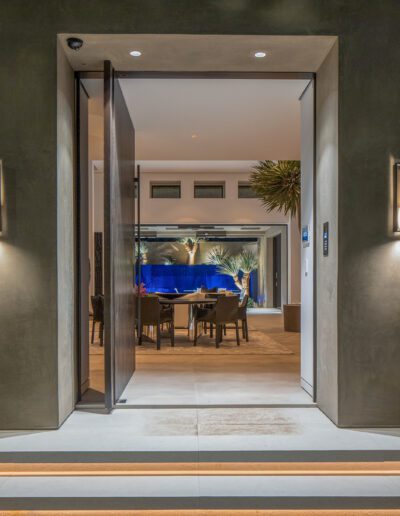 An illuminated modern home entrance at dusk, showcasing an open door leading to a stylish interior with a dining area visible in the background.