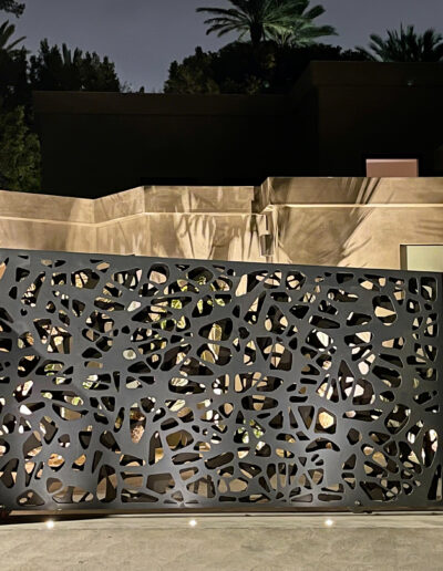 A decorative metal gate with cut-out patterns illuminated at night in front of a modern residence.