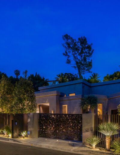 Modern house with landscape lighting at twilight, surrounded by a metal fence and lush vegetation.