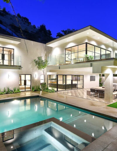 Modern two-story house with abundant lighting, a balcony, and a pool at twilight.